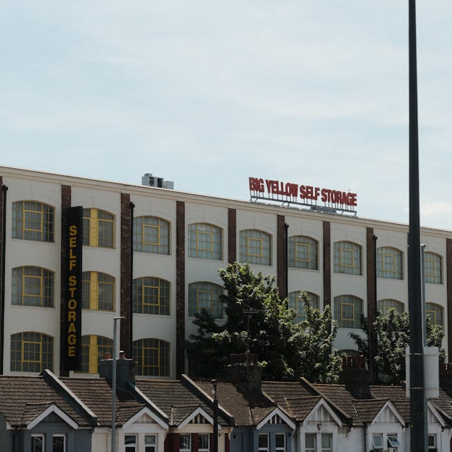 Exterior view of a multi-storey commercial building with a sign reading 'Big Yellow Self Storage' in red letters at the top. The building features large, yellow-framed windows arranged in a grid pattern, with white walls and vertical brick accents. A vertical yellow sign on the side of the building displays 'Self Storage' in black letters. In front of the building, there are small residential houses with sloped roofs and chimneys, casting shadows on the pavement. Several trees partially obscure the lower part of the building, and a lamppost is visible on the right side. The scene is captured on a clear day with partly cloudy skies, highlighting the building's role in providing storage solutions, relevant to house removals and packing services offered by companies like Man with Van Finchley.