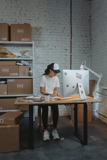 A woman wearing casual clothing, including a white t-shirt, dark pants, white sneakers, and a cap, is standing next to a wooden desk inside an industrial-style room with white brick walls. She appears to be packing or preparing documents, with various papers, a calculator, and a coffee mug on the desk. Behind her, there is a white shelving unit filled with cardboard boxes, some opened, and sealed with packing labels, indicating storage or preparations for a home or office move. To her right, a large white appliance or piece of equipment is positioned near a window, with a white desk lamp providing additional lighting. The scene is well-lit, with natural and artificial light highlighting the workspace. This setup suggests an environment related to packing or organizing belongings for a furniture transport or house removal service, aligning with professional moving or relocation activities conducted by Man with Van Finchley, as part of a small business move or home relocation process.
