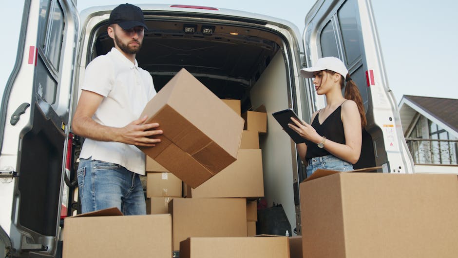 A man with a beard, wearing a white polo shirt, jeans, and a black cap, is inside the open rear of a white moving van, lifting and carrying a large cardboard box. Next to him, a woman with long brown hair, wearing a white cap, black sleeveless top, and light blue jeans, is standing outside the vehicle, holding a tablet device and observing the loading process. The van’s interior is filled with multiple cardboard boxes of various sizes, some stacked on each other, prepared for a home relocation. The scene is set outdoors, with a residential house visible in the background under a clear sky. Moving equipment such as straps or blankets are not visible, but the conveying of packing and furniture transport is implied by the scene. The image illustrates the loading process involved in house removals, ensuring careful handling of packed items for a professional move, as represented by Man with Van Finchley.