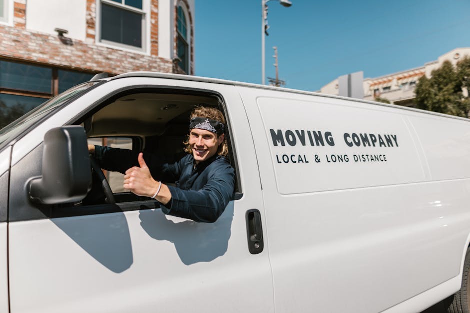 A smiling young man with shoulder-length hair, wearing a headband and dark clothing, is sitting in the driver's seat of a white moving van. His arm is extended out of the window, giving a thumbs-up gesture. The van has bold black lettering indicating it is part of a moving company specializing in local and long-distance relocations, with the text 'MOVING COMPANY' and 'LOCAL & LONG DISTANCE'. The vehicle is parked outdoors in an urban environment, with a clear blue sky overhead. In the background, there are modern buildings with glass windows, a streetlight, and some greenery. The scene captures the loading process or preparation phase of a house removal, with the focus on the vehicle and the driver engaged in home relocation activities, supported by [COMPANY_NAME] for furniture transport and packing and moving services.