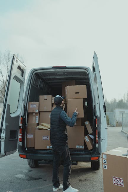 A man with a beard, wearing a white polo shirt, jeans, and a black cap, is inside the open rear of a white moving van, lifting and carrying a large cardboard box. Next to him, a woman with long brown hair, wearing a white cap, black sleeveless top, and light blue jeans, is standing outside the vehicle, holding a tablet device and observing the loading process. The van’s interior is filled with multiple cardboard boxes of various sizes, some stacked on each other, prepared for a home relocation. The scene is set outdoors, with a residential house visible in the background under a clear sky. Moving equipment such as straps or blankets are not visible, but the conveying of packing and furniture transport is implied by the scene. The image illustrates the loading process involved in house removals, ensuring careful handling of packed items for a professional move, as represented by Man with Van Finchley.