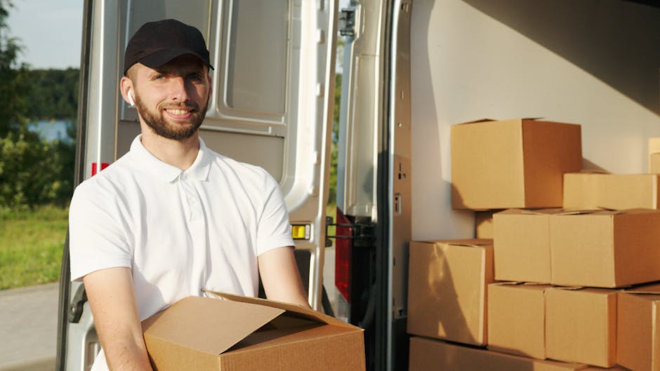 A man with light skin, a beard, and wearing a black cap and white polo shirt is standing outside near a white moving van during a home relocation process. He is holding a cardboard box, which appears to be packed for a furniture transport or packing and moving service. The side door of the van is open, revealing a stack of cardboard boxes inside, some placed inside the vehicle and others on the pavement nearby. The boxes vary in size and are sealed with packing tape. The van is parked on a paved area adjacent to a grassy landscape with trees and a body of water in the background, under clear daylight. Visible equipment includes a trolley or dolly, used for lifting and transporting boxes during the loading process. The scene depicts the careful handling and loading of packed items typical of professional removals services, aligning with the context of house removals and moving logistics provided by Man with Van Finchley.