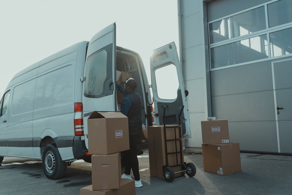 A professional mover from Man with Van Finchley is loading packed cardboard boxes and a black hand truck onto a white panel van parked outside a modern building with large glass windows. The mover, dressed in casual work attire with a vest and cap, is seen placing a box inside the vehicle, which has its rear doors wide open, revealing more boxes and packing materials inside. Several additional cardboard boxes are positioned on the pavement nearby, some stacked and others waiting to be loaded. The scene is set during daylight, with natural sunlight creating a slightly bright atmosphere, emphasizing the moving logistics involved in house relocation and furniture transport. The environment appears clean and organized, with the van ready for transport as part of a comprehensive packing and moving service typically provided by companies like Man with Van Finchley, aimed at facilitating efficient home removals and logistical support for first-time renters and homeowners alike.