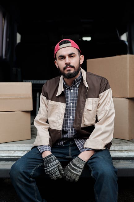 A man with light skin, a beard, and wearing a black cap and white polo shirt is standing outside near a white moving van during a home relocation process. He is holding a cardboard box, which appears to be packed for a furniture transport or packing and moving service. The side door of the van is open, revealing a stack of cardboard boxes inside, some placed inside the vehicle and others on the pavement nearby. The boxes vary in size and are sealed with packing tape. The van is parked on a paved area adjacent to a grassy landscape with trees and a body of water in the background, under clear daylight. Visible equipment includes a trolley or dolly, used for lifting and transporting boxes during the loading process. The scene depicts the careful handling and loading of packed items typical of professional removals services, aligning with the context of house removals and moving logistics provided by Man with Van Finchley.
