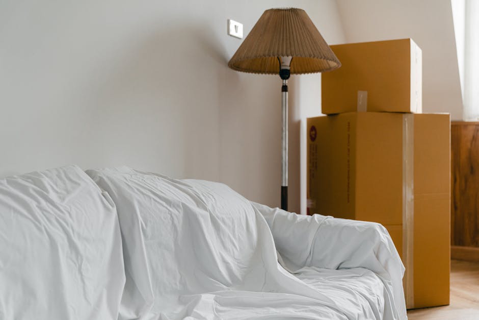 Image of a room during relocation preparations, featuring a white fabric-covered sofa positioned against a light beige wall. Next to the sofa, there is a wooden floor lamp with a brown pleated lampshade, standing beside several large cardboard moving boxes stacked vertically near the corner of the room. The boxes are sealed with packing tape, and some have printed labels or barcodes visible. The space is illuminated by natural light coming through a window, partially visible on the right side, casting soft shadows within the room. This setting illustrates the typical packing and home relocation process, with furniture and items ready for transport. The scene is captured inside a residential interior, highlighting elements associated with house removals, and subtly referencing the services offered by Man with Van Finchley for furniture transport and packing logistics, as suggested by the context of the page about quick moves in Finchley.