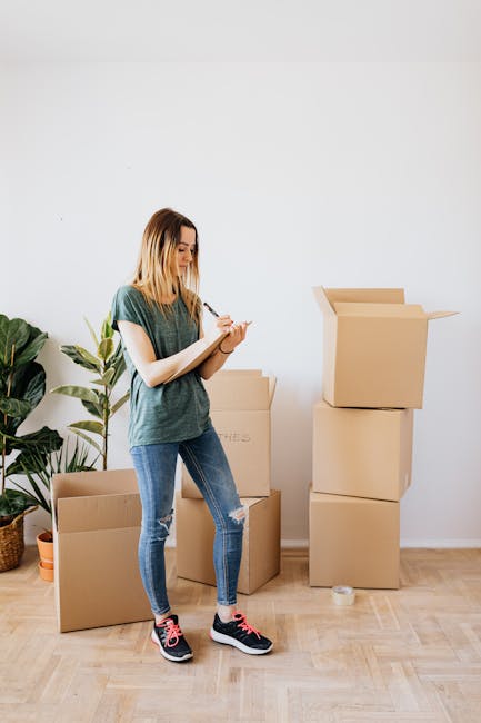 A young woman with shoulder-length blonde hair, dressed in a casual green t-shirt, ripped blue jeans, and black sneakers with pink accents, is standing indoors against a plain white wall. She is holding a clipboard and pen, appearing to be making notes or checking a list. Surrounding her are several cardboard moving boxes of varying sizes, some of which are open or partially closed, with packing paper and tape visible. A large potted plant with broad green leaves is positioned to her left, adding a touch of greenery to the scene. The boxes are arranged on a light wooden floor, indicating an ongoing home relocation or packing process. This setting reflects the activities of packing and organizing belongings, typical of furniture transport and moving logistics services offered by Man with Van Finchley, as part of their house removals solutions for quick moves in the Finchley area.